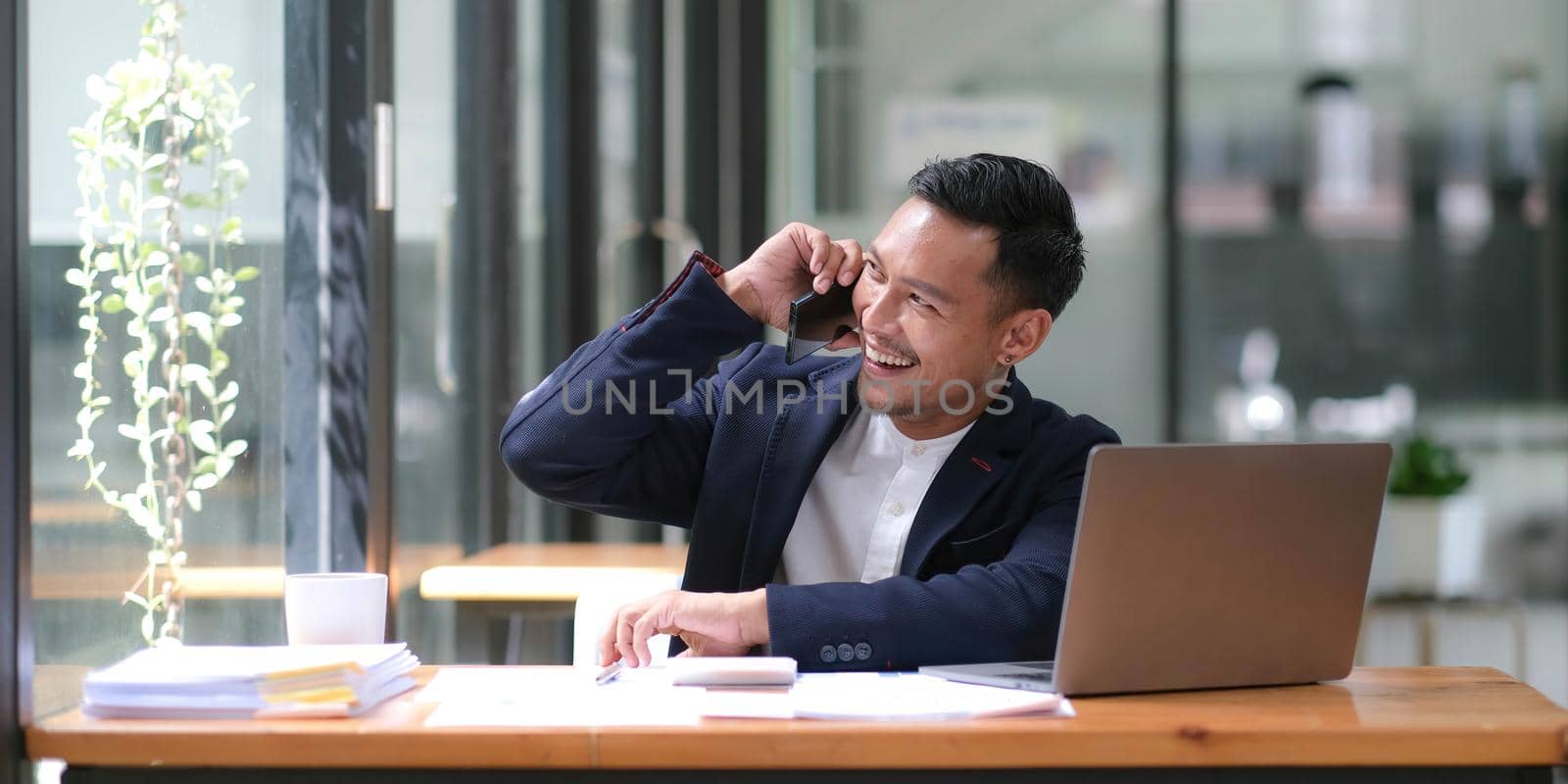 Asian businessman sitting on the phone with a customer with a laptop and document at his office desk..