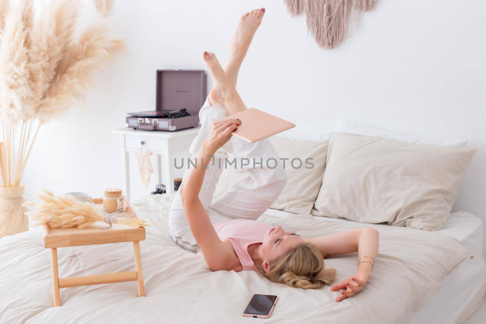 A beautiful woman with blond hair, in a pink top and white trousers, lies on her back on a white bed in the morning, legs raised, looks into a pink digital tablet.