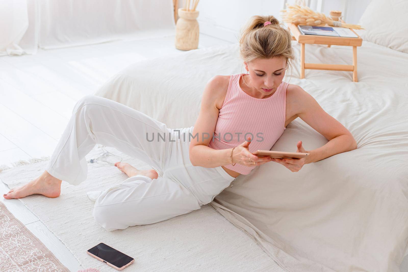 A beautiful slender woman with blond hair, a pink top and white pants, sits on the floor in the morning, near a white bed, hold a digital tablet, a smartphone lies nearby