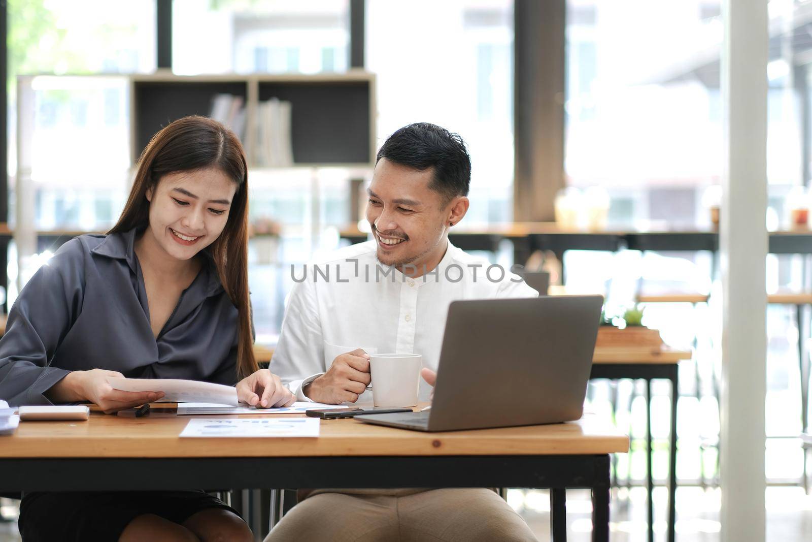 Two employees in a modern office, an Asian man and a woman working at a table, colleagues discussing and consulting, thinking about a joint project by wichayada