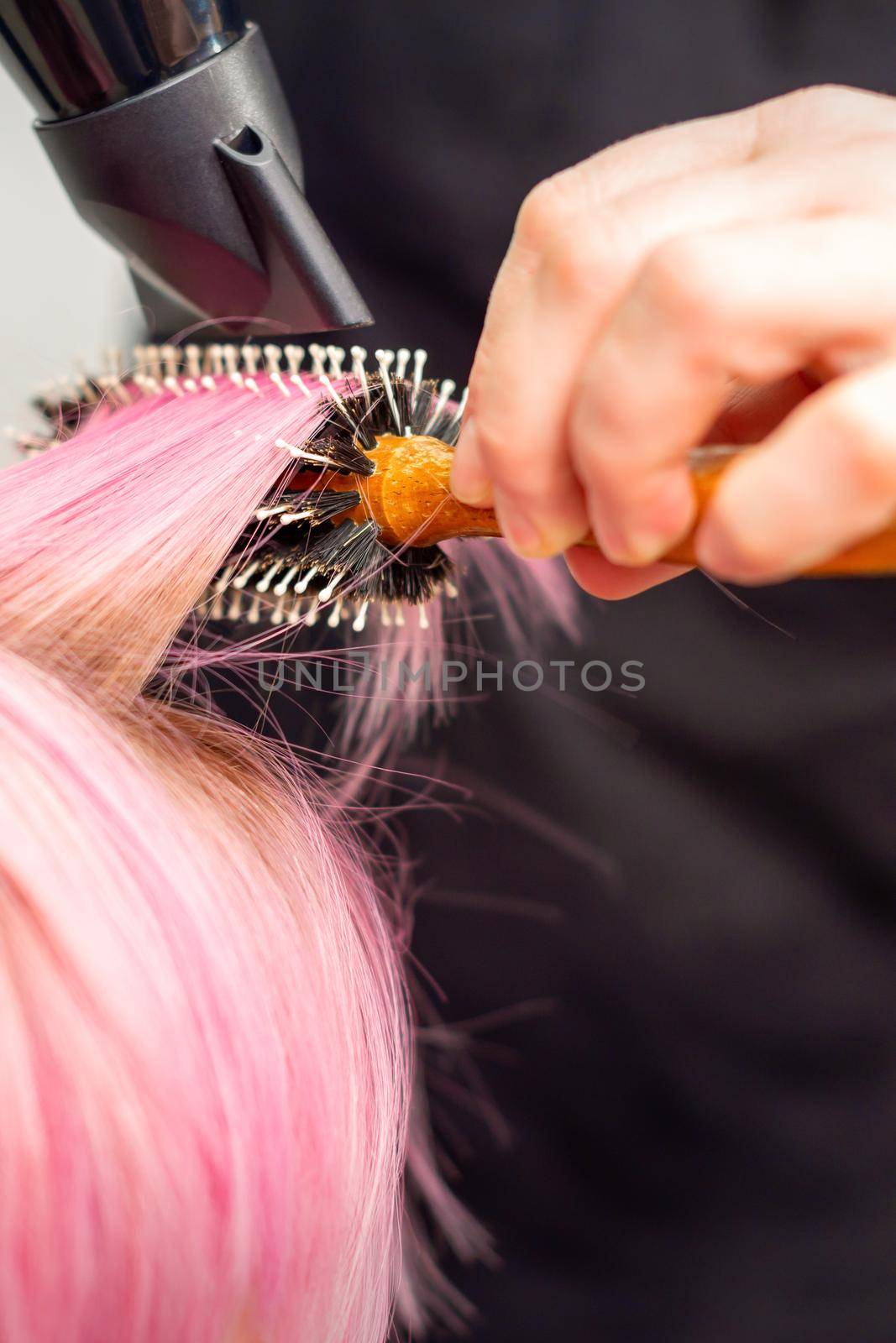Drying short pink hair of young caucasian woman with a black hairdryer and black round brush by hands of a male hairdresser in a hair salon, close up. by okskukuruza