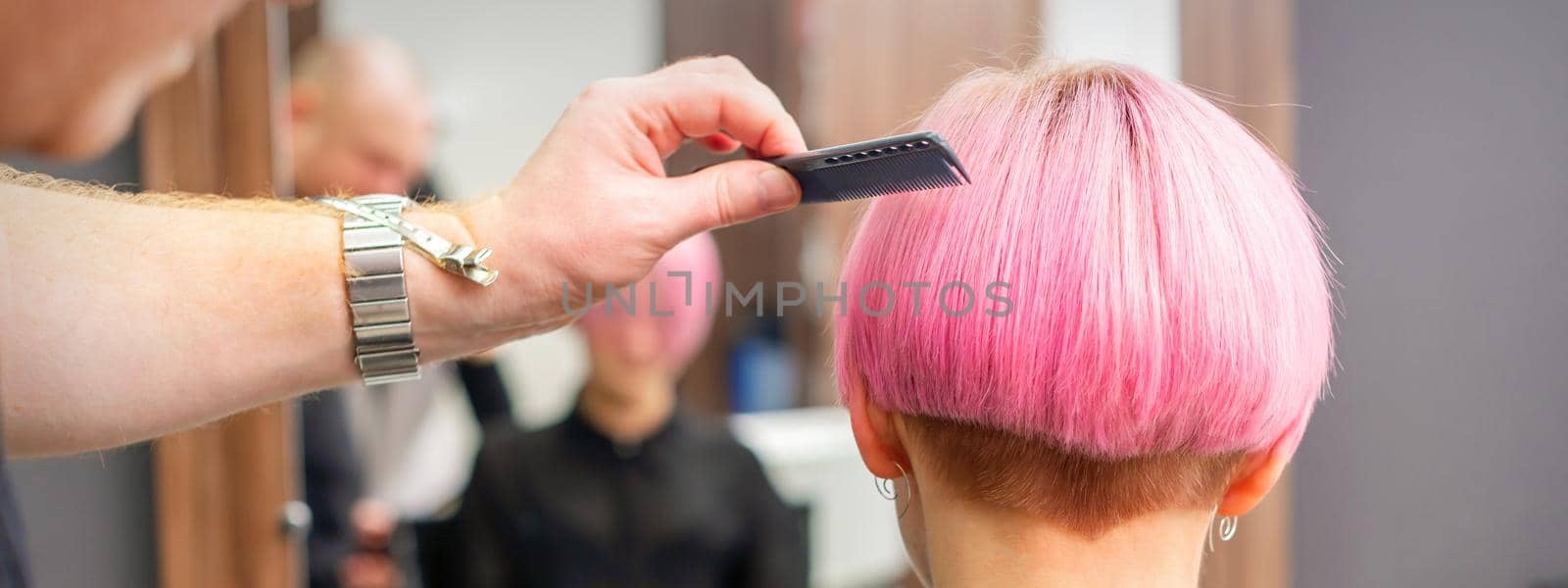 A hairdresser is combing the dyed pink short hair of the female client in hairdresser salon, back view