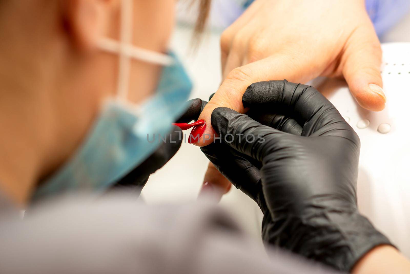 Manicure varnish painting. Close-up of a manicure master wearing rubber black gloves applying red varnish on a female fingernail in the beauty salon. by okskukuruza