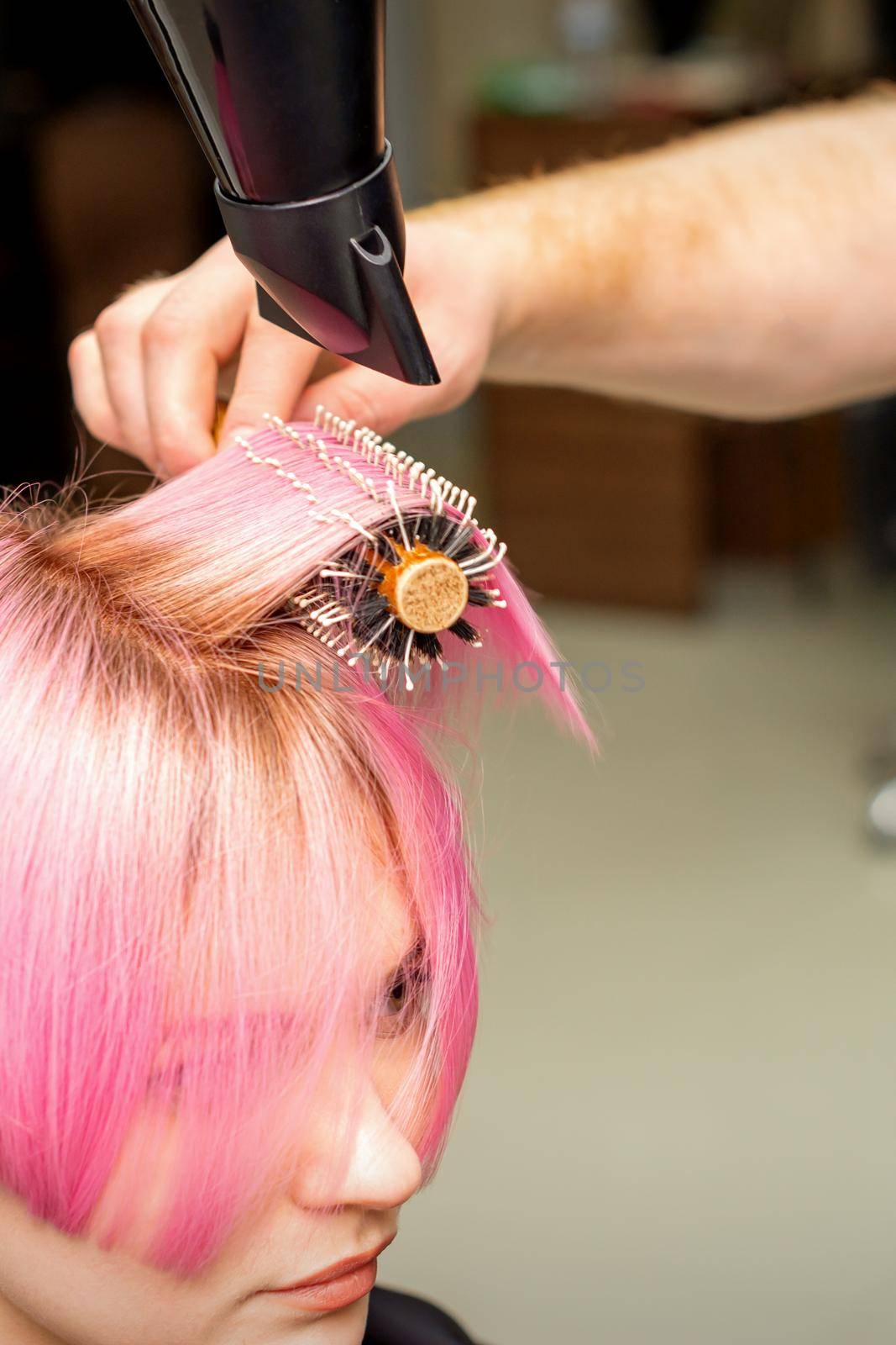 Drying short pink hair of young caucasian woman with a black hairdryer and black round brush by hands of a male hairdresser in a hair salon, close up. by okskukuruza