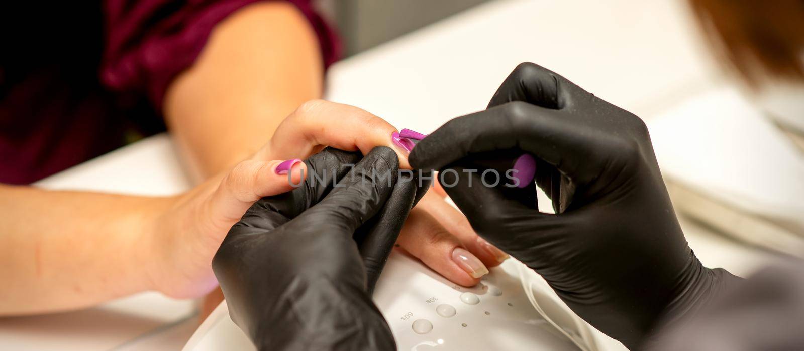 Professional manicure. A manicurist is painting the female nails of a client with purple nail polish in a beauty salon, close up. Beauty industry concept. by okskukuruza