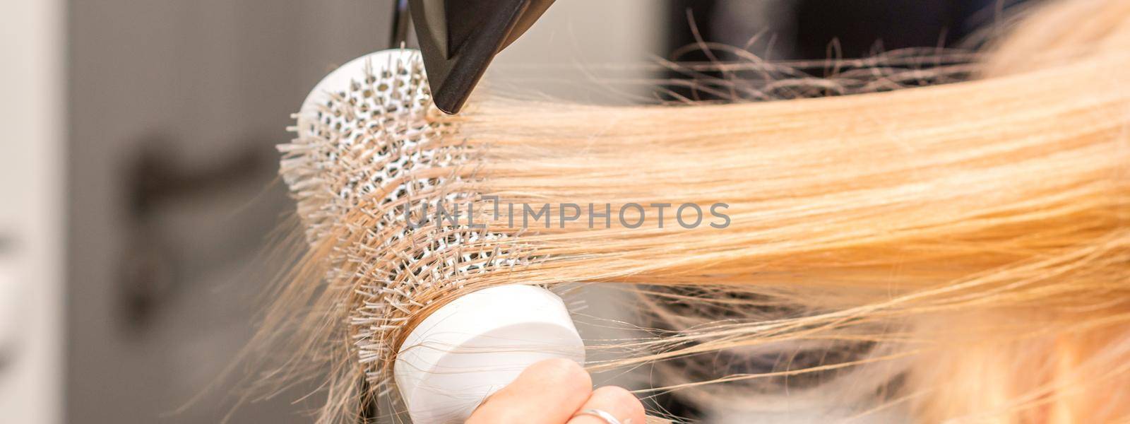 Drying straight blond hair with black hairdryer and white round brush in hairdresser salon, close up. by okskukuruza