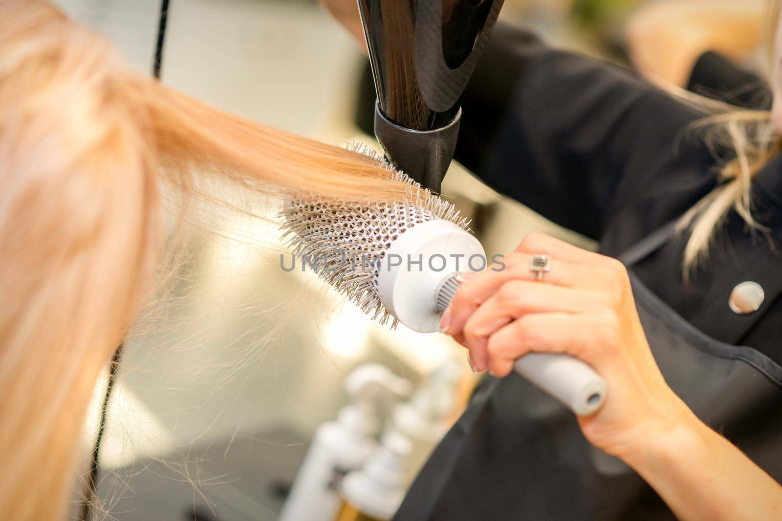 Drying straight blond hair with black hairdryer and white round brush in hairdresser salon, close up. by okskukuruza