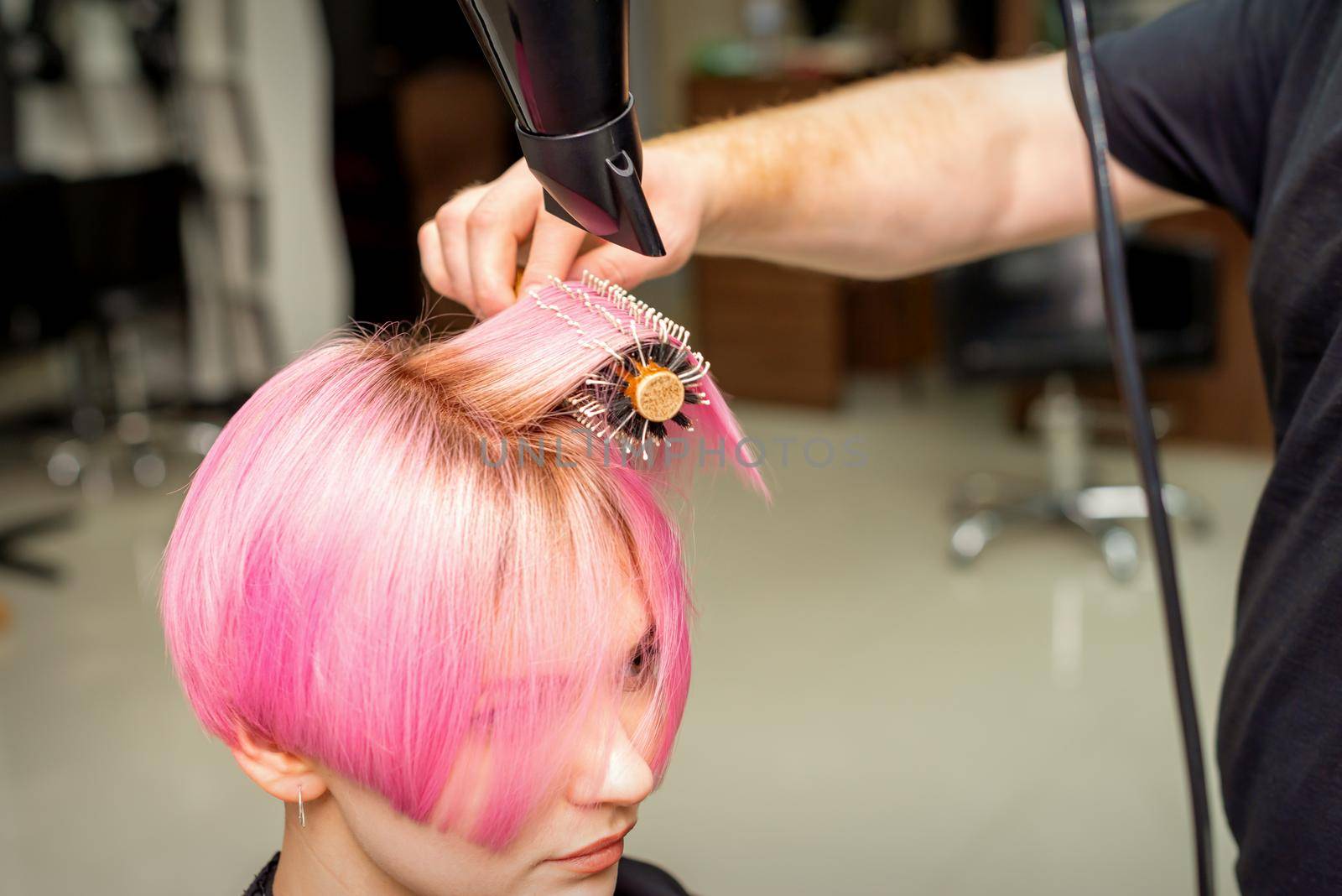 Drying short pink hair of young caucasian woman with a black hairdryer and black round brush by hands of a male hairdresser in a hair salon, close up