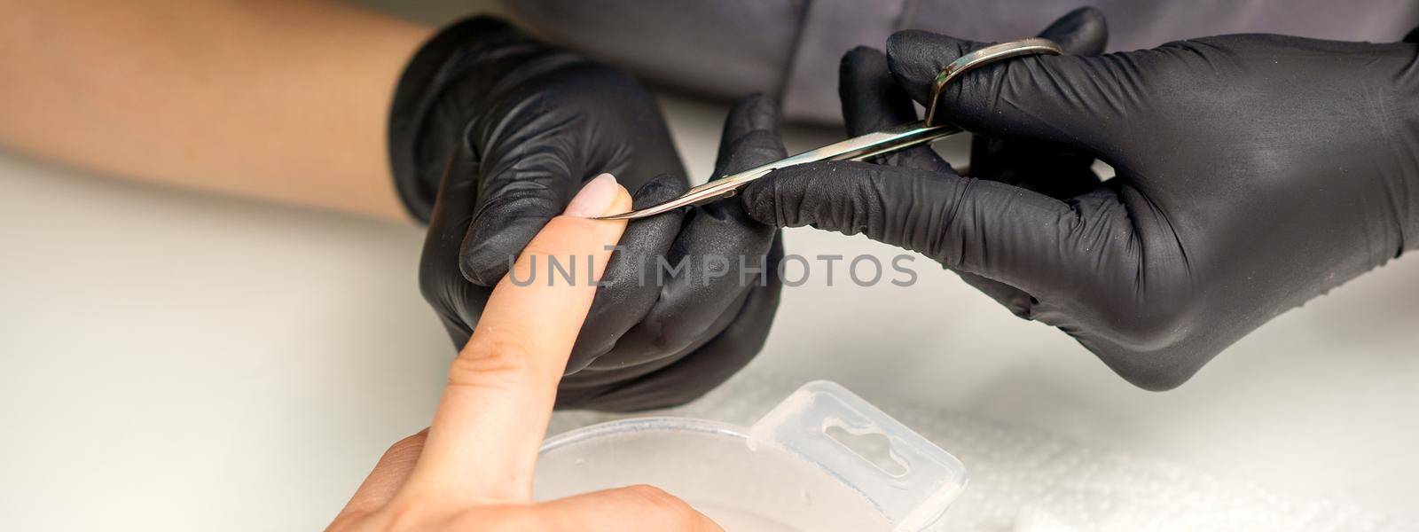 Manicure master removes cuticles from female nails with scissors wearing protective gloves in manicure salon. by okskukuruza