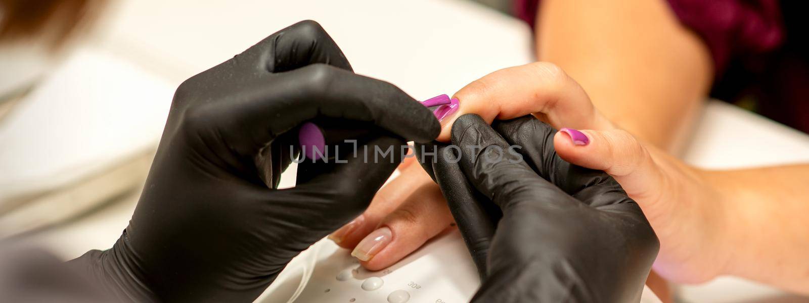 Professional manicure. A manicurist is painting the female nails of a client with purple nail polish in a beauty salon, close up. Beauty industry concept. by okskukuruza