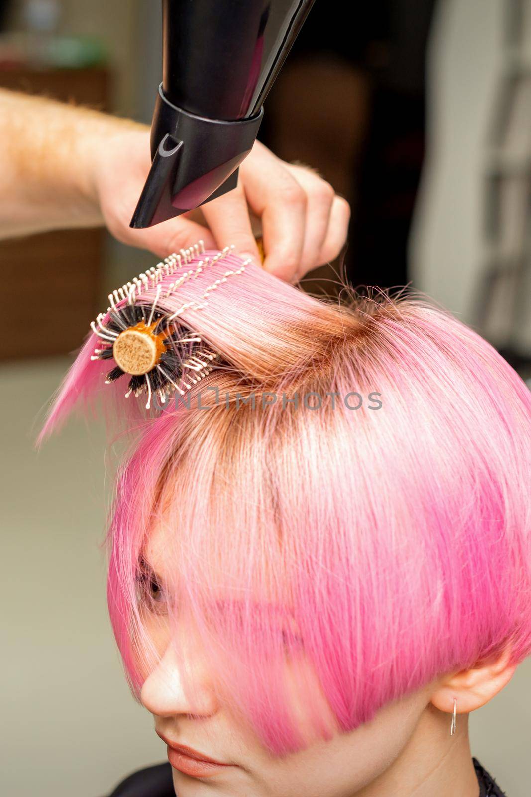 Drying short pink hair of young caucasian woman with a black hairdryer and black round brush by hands of a male hairdresser in a hair salon, close up. by okskukuruza