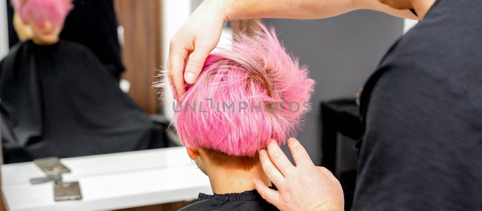 Hairdresser with hands is checking out and fixing the short pink hairstyle of the young white woman in a hair salon. by okskukuruza