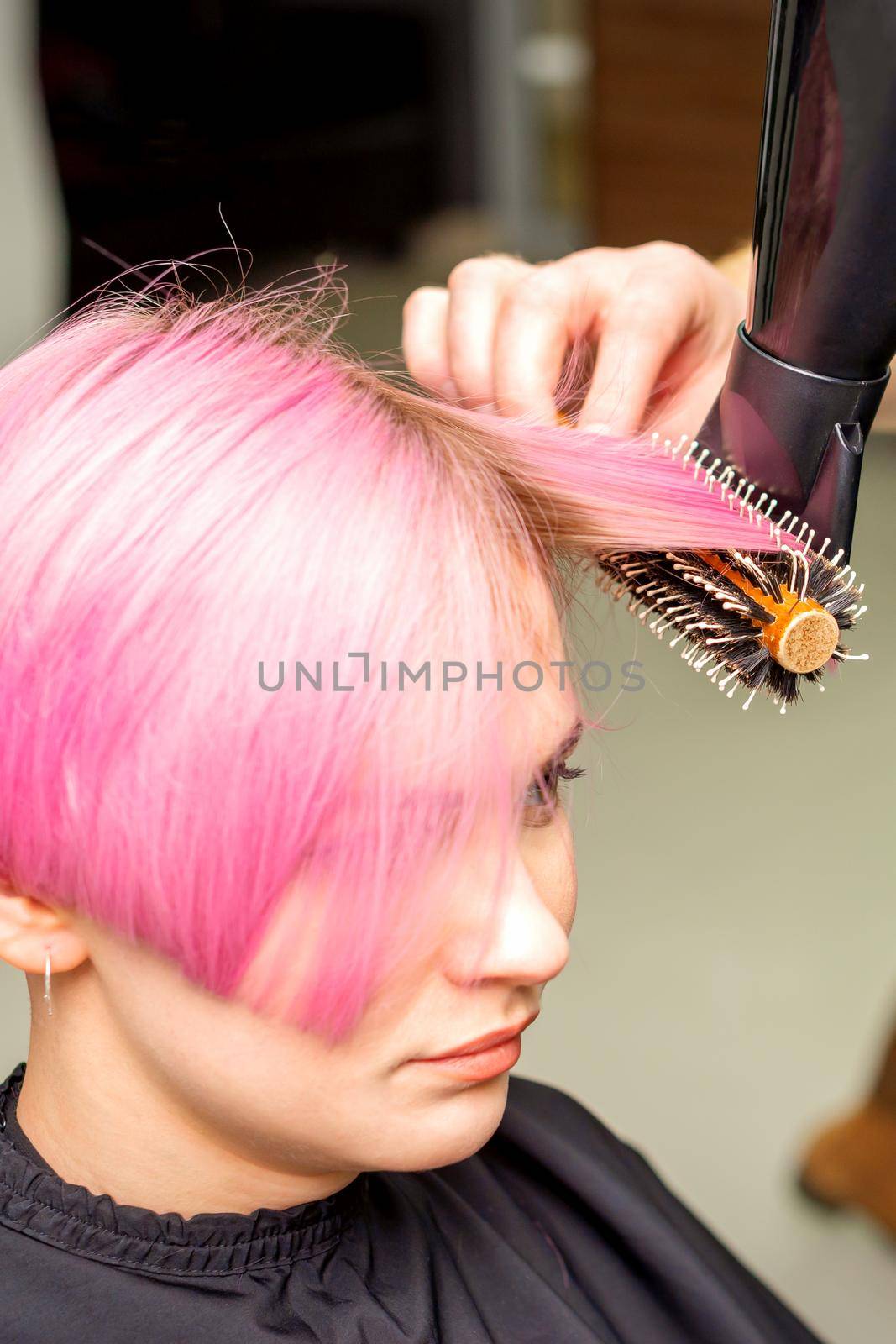 Drying short pink hair of young caucasian woman with a black hairdryer and black round brush by hands of a male hairdresser in a hair salon, close up