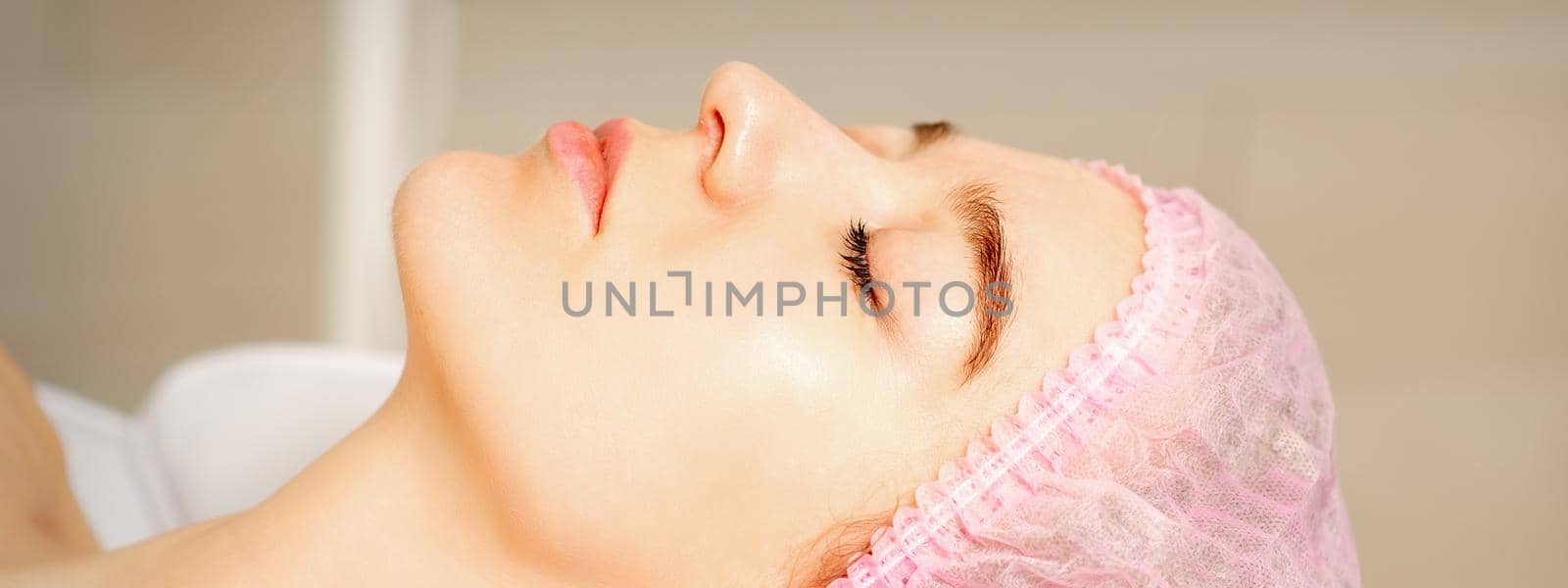 Side view of a young caucasian woman in a medical cap lying with closed eyes waiting for the cosmetic procedure in a beauty salon. by okskukuruza