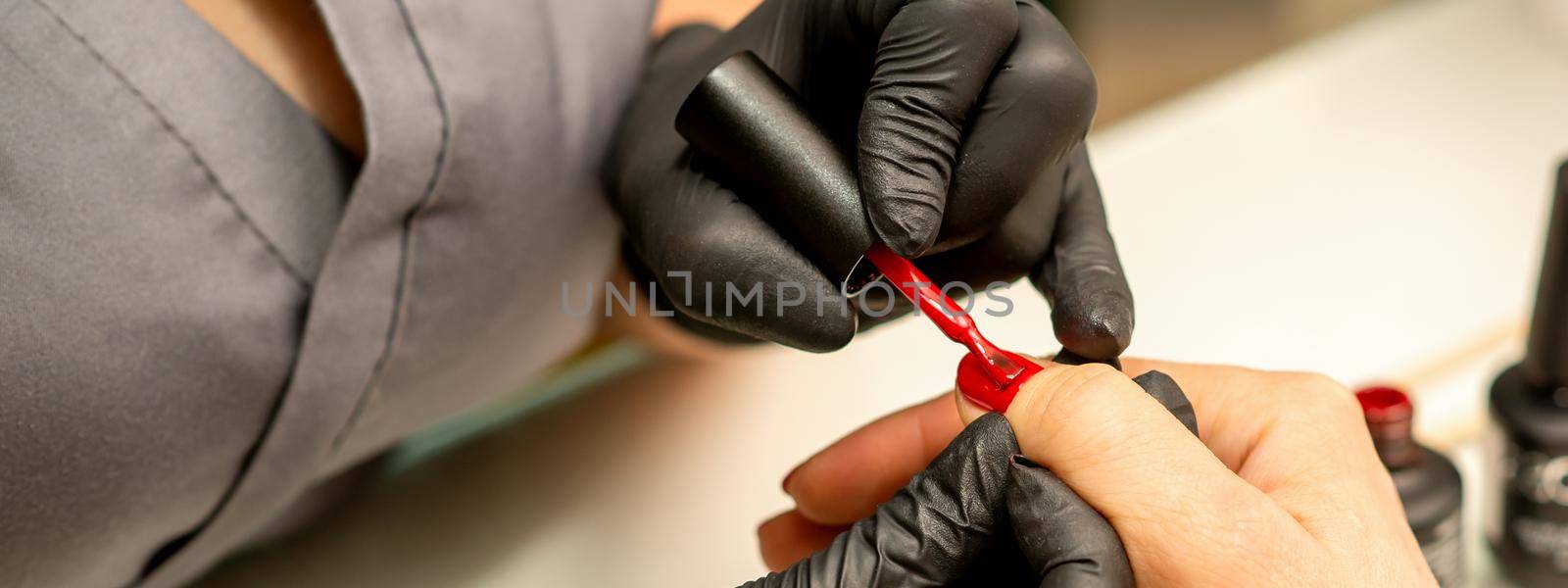 Professional manicure. A manicurist is painting the female nails of a client with red nail polish in a beauty salon, close up. Beauty industry concept