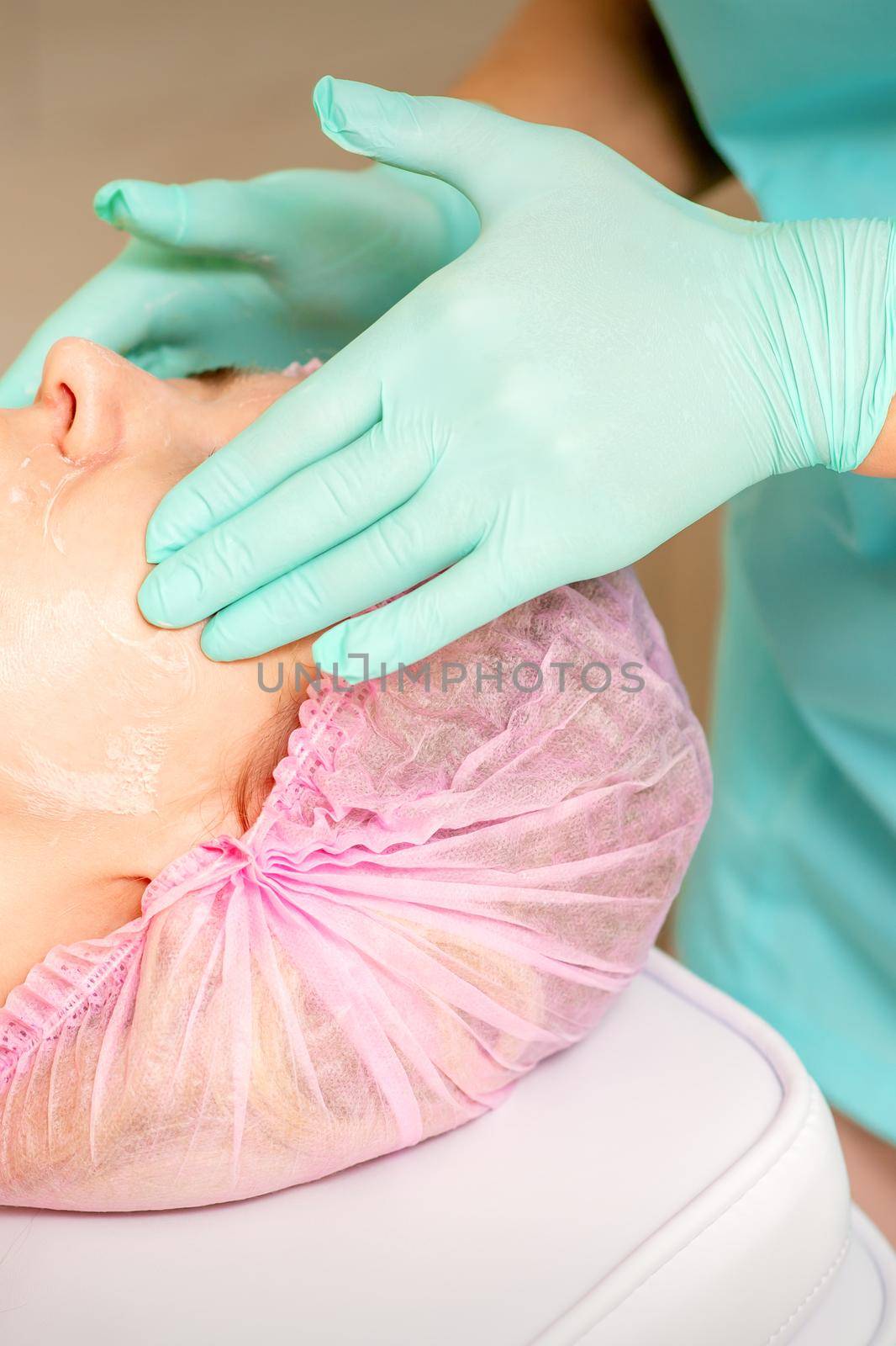 Cosmetologist with gloved hands applies a moisturizing mask with peeling cream on the female face. Facial cosmetology treatment. Procedures for facial care