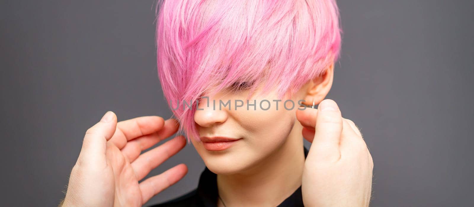 Hairdresser with hands is checking out and fixing the short pink hairstyle of the young white woman in a hair salon. by okskukuruza