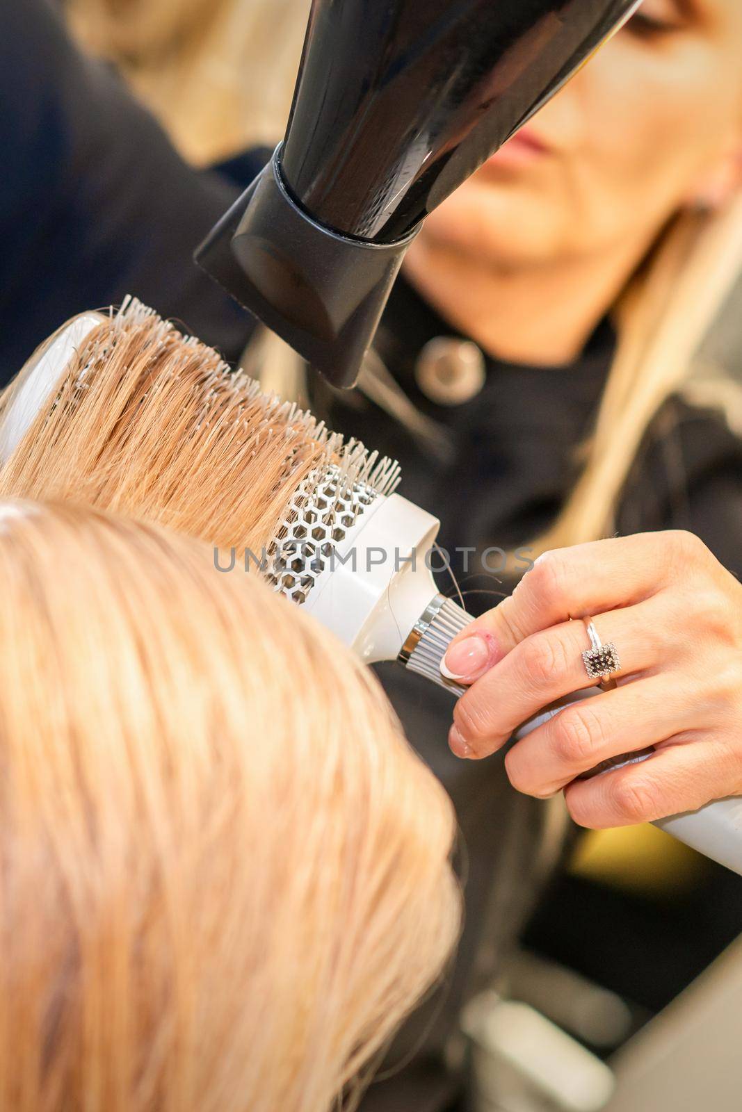 Drying straight blond hair with black hairdryer and white round brush in hairdresser salon, close up