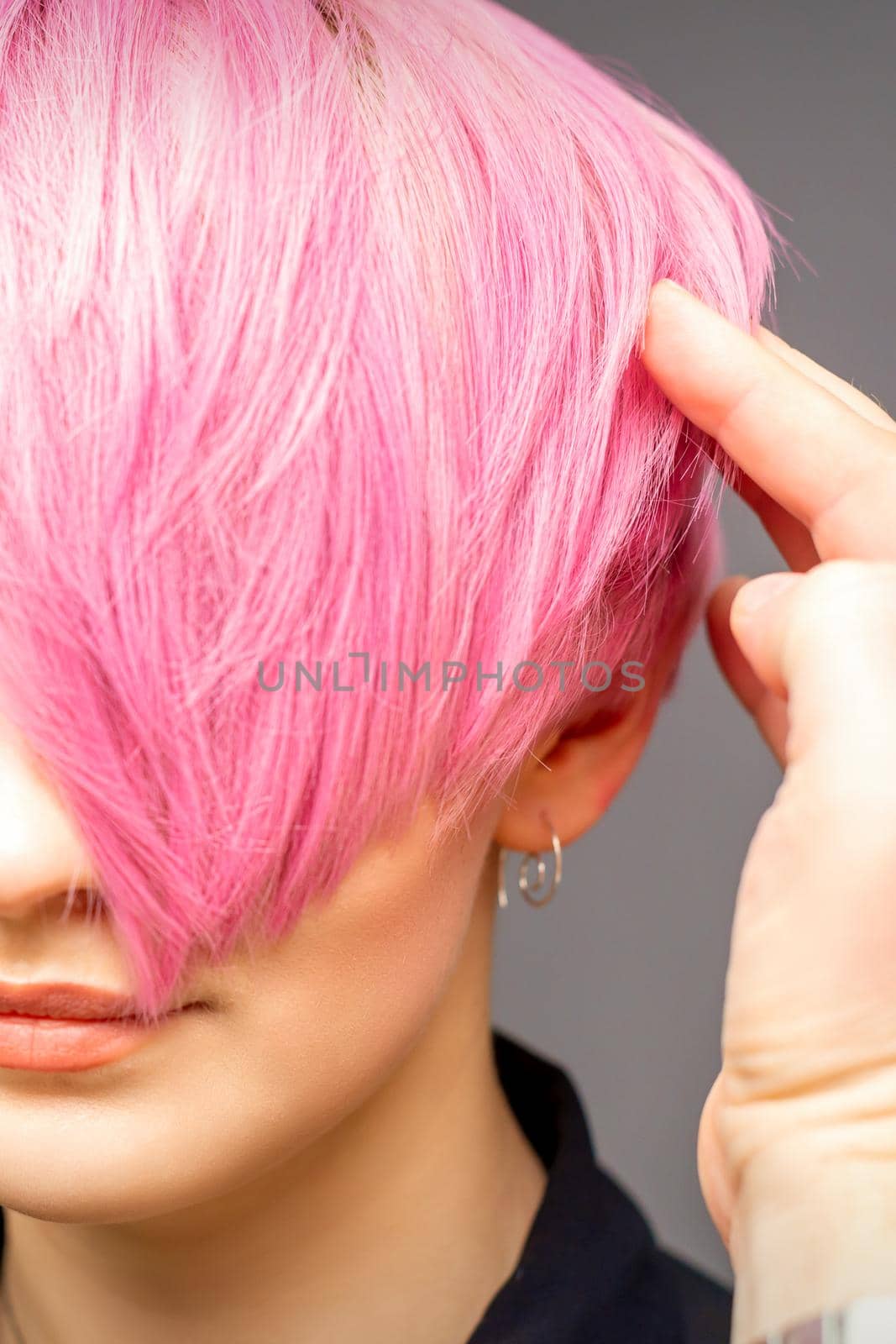 Hairdresser with hands and comb is checking out and fixing the short pink hairstyle of the young white woman in a hair salon. by okskukuruza