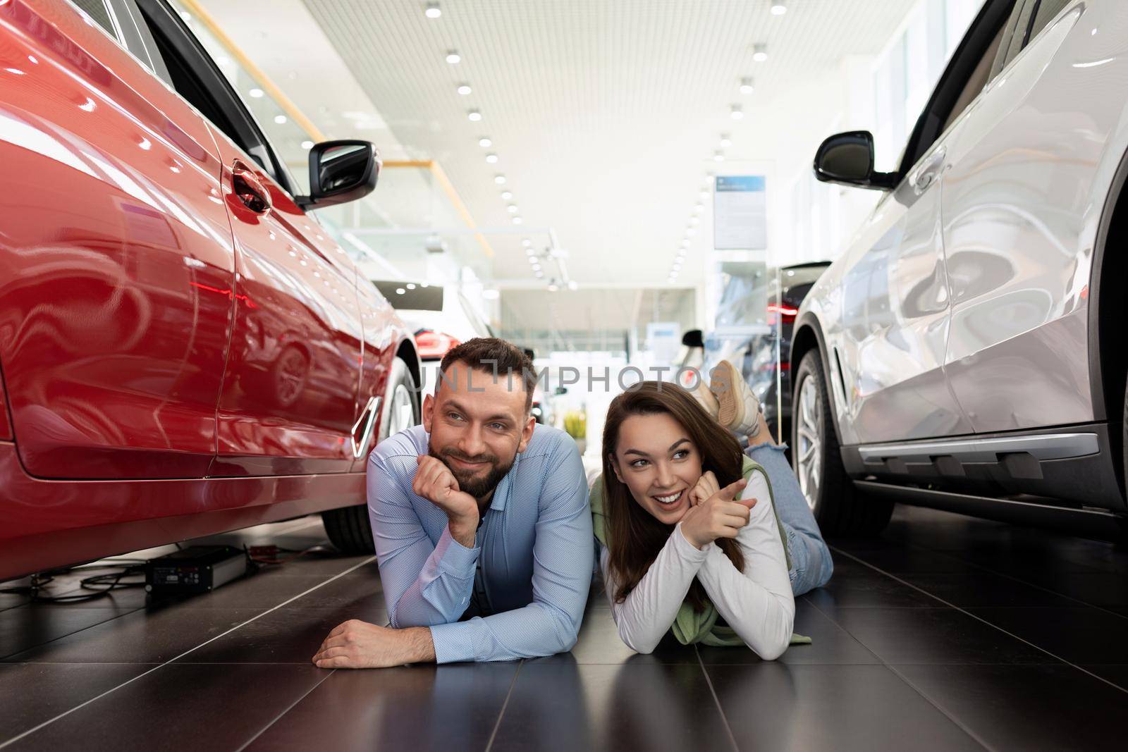 cheerful young couple fooling around lies on the floor in a car dealership dreaming about a new car by TRMK