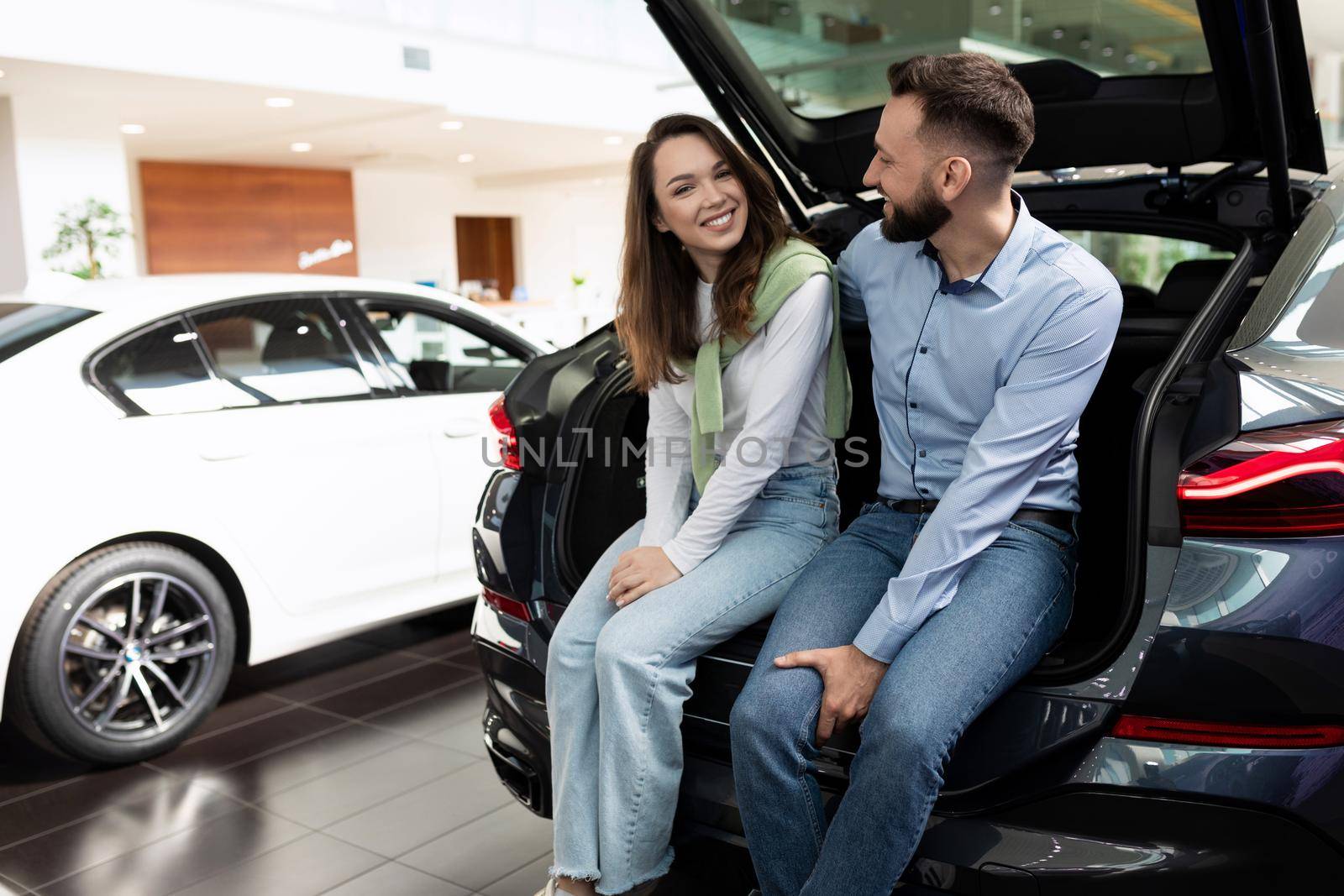 a young married couple in a car dealership chooses a car sits with a smile in the trunk of a crossover.