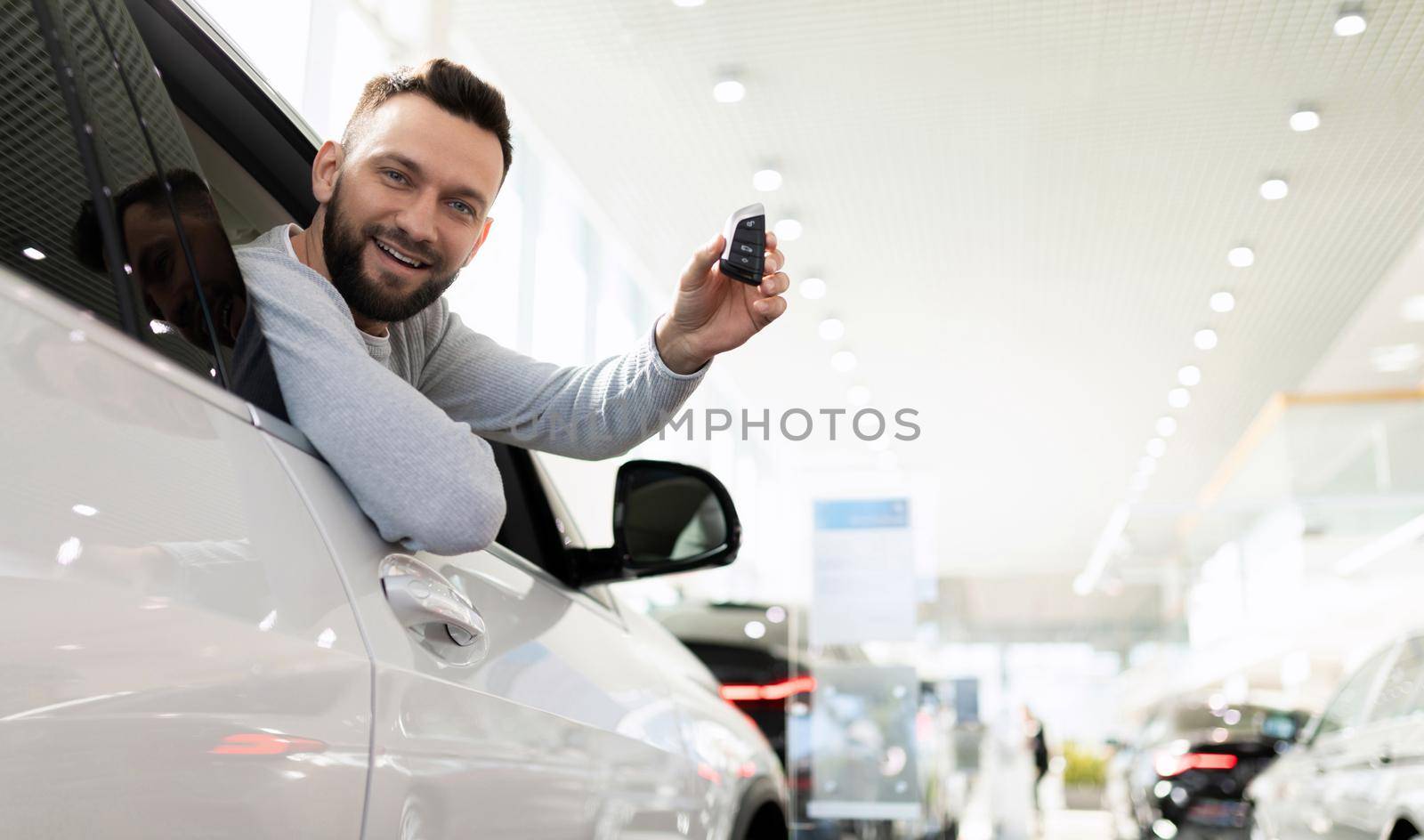 happy owner of a new car with a key in his hands in a dealership showroom looks out of the car.
