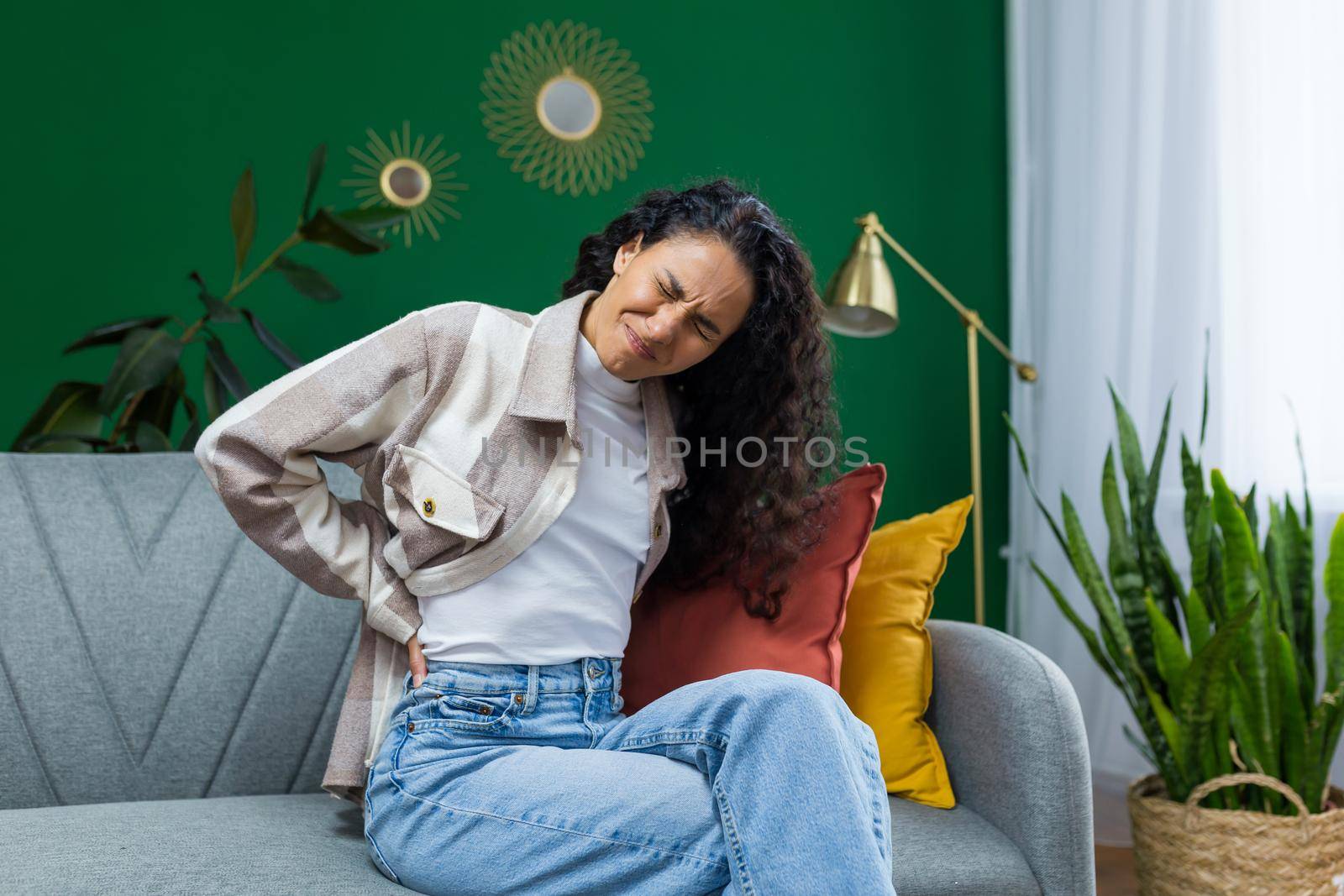 Young beautiful hispanic woman at home having severe back pain, housewife sitting on sofa in living room and massaging back side in room with green wall.