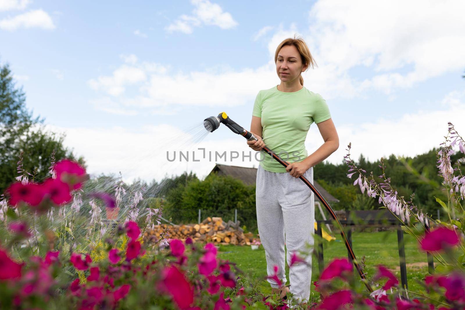 a young woman with a smile waters a flower bed from a garden hose with a sprayer.