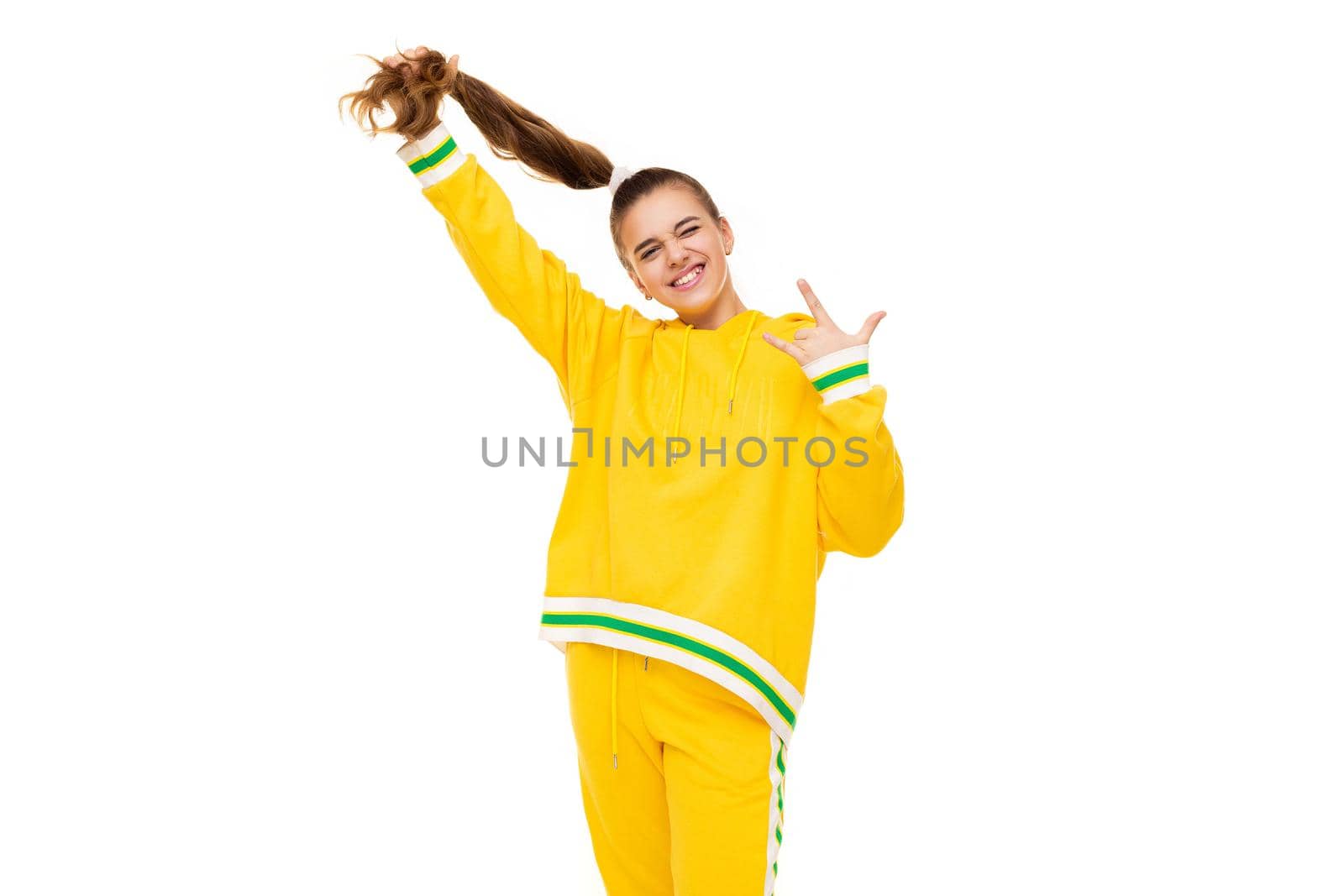 Photo of a beautiful smiling girl with dark hair with a ponytail in a stylish yellow tracksuit with green stripes shows rock hand isolated on white background with free space for text by TRMK