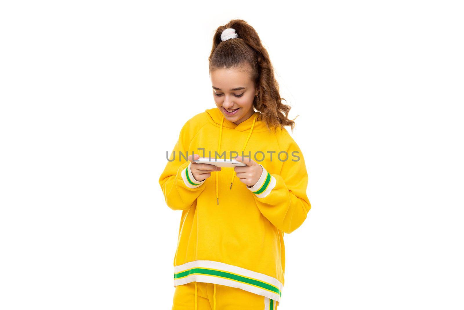 Pretty beautiful smiling cheerful cute girl with dark hair with a ponytail in a stylish yellow tracksuit with green stripes plays a mobile phone with a white case isolated on a white background with free space for text.