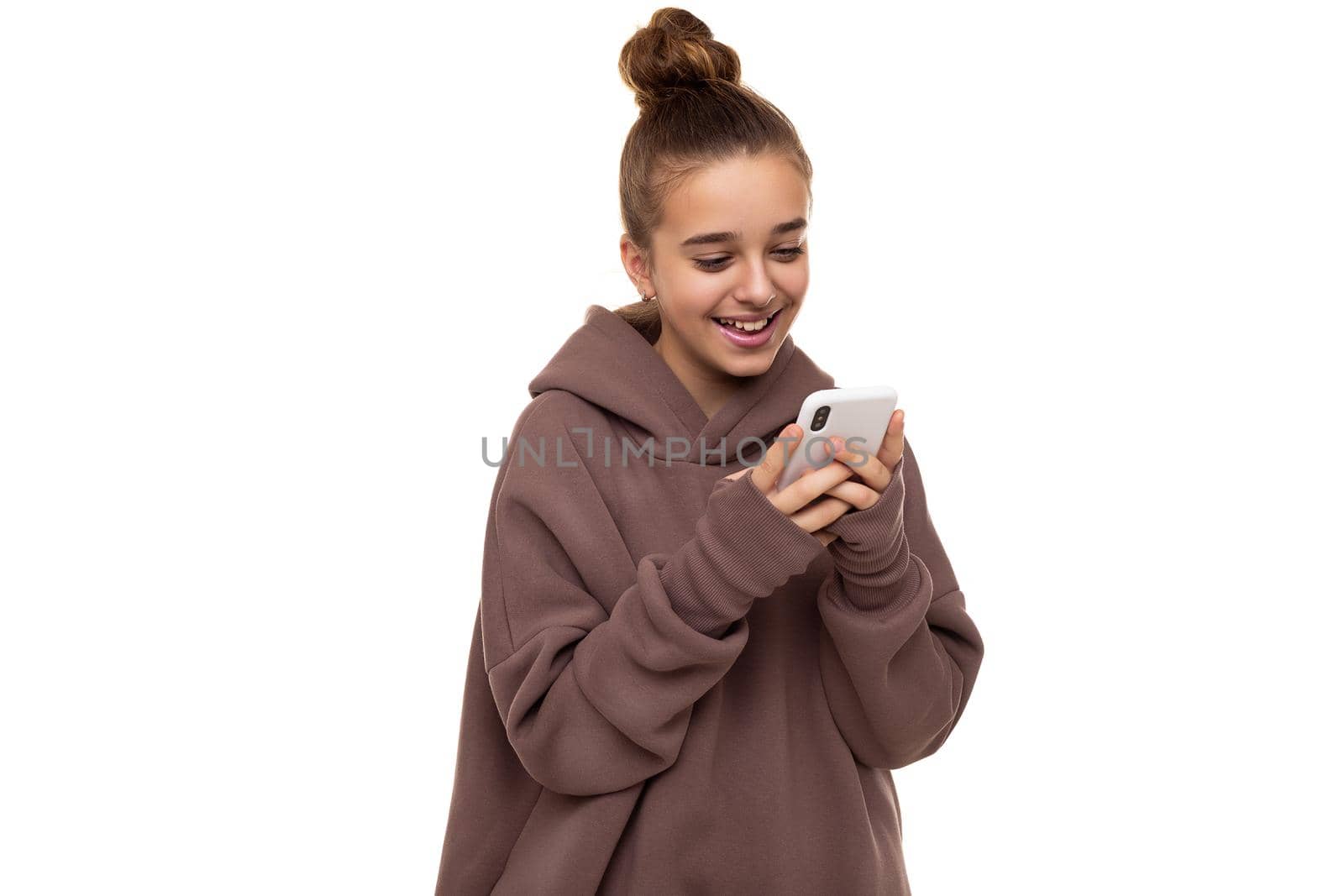 Photo of a beautiful positive cute girl with dark hair and a bun in a brown hoodie with a hood holding a mobile phone and texting in social networks isolated on a white background with free space for text.