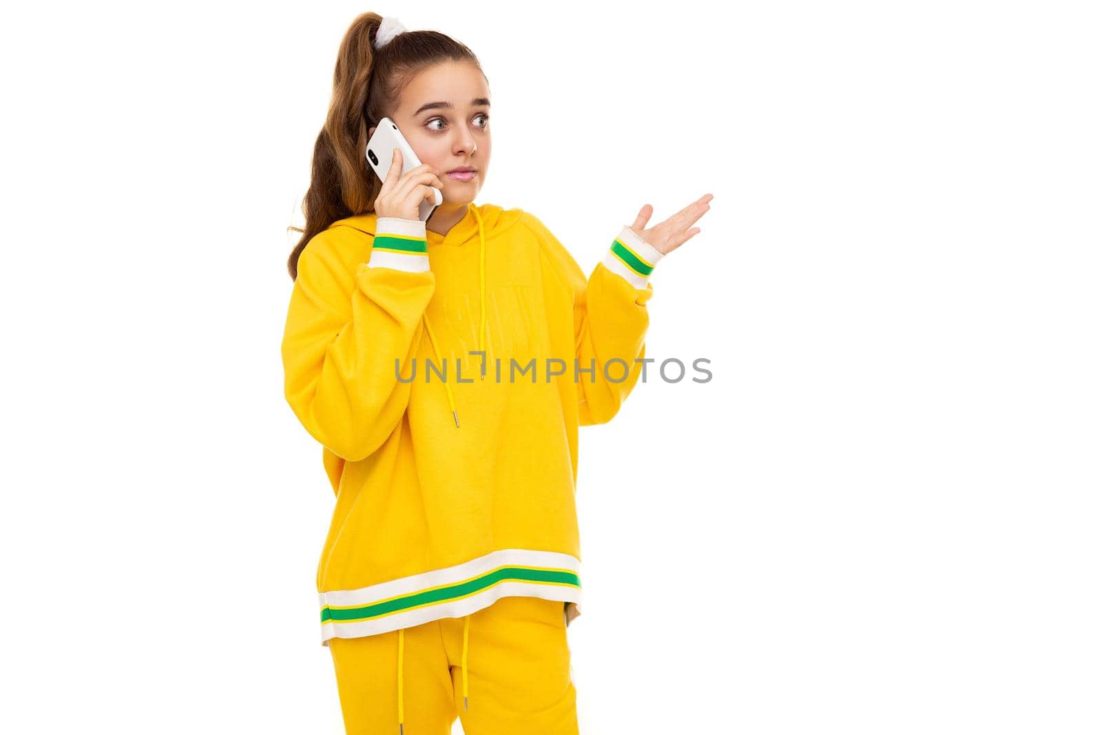 Photo of a beautiful shocked and surprised girl with dark hair with a ponytail in a stylish yellow tracksuit with green stripes talking on a mobile phone isolated on white background with free space for text.