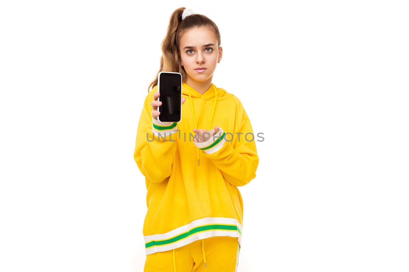 Photo of a beautiful girl with dark hair with a ponytail in a stylish yellow tracksuit with green stripes shows a mobile phone with a black blank display isolated on a white background with free space for text by TRMK