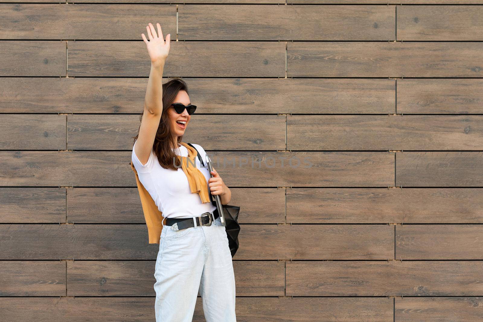 Photo of fascinating beautiful smiling charming young brunette girl wearing stylish clothes holding computer laptop and black sunglasses in white t-shirt and light blue jeans in the street near brown wall and waving with hand.