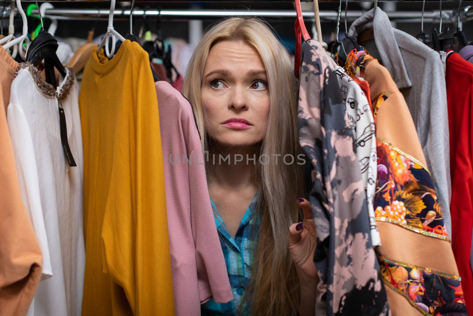 Girl worried because she hasn't found the dress she dreamed of. A blonde girl among hangers of clothes with a grim face.