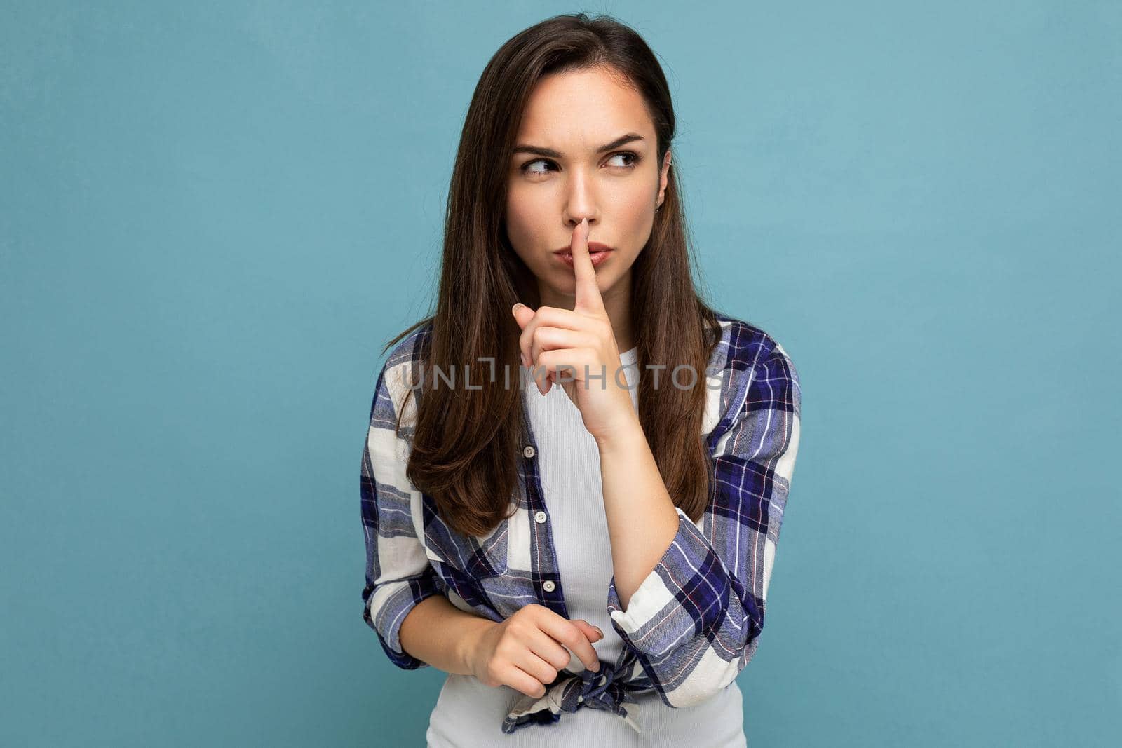 Young serious angry beautiful brunette woman with sincere emotions wearing trendy check shirt standing isolated on blue background with empty space for text and showing shh gesture. Keep secret concept.