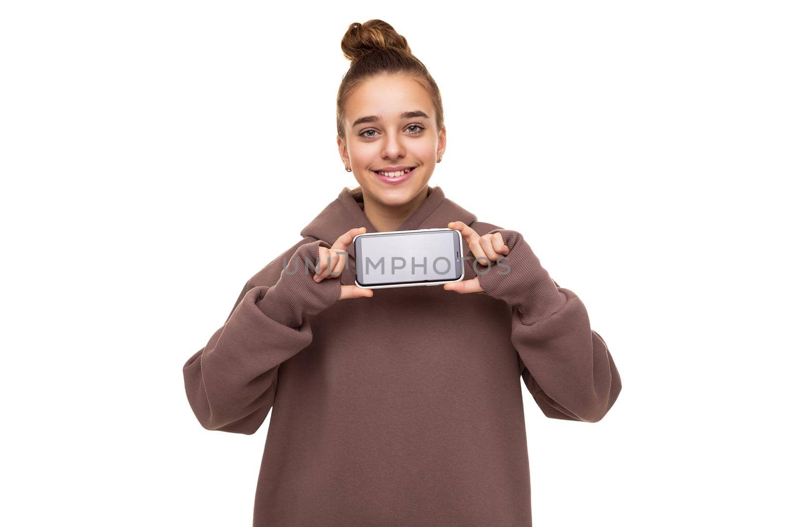 Photo of a beautiful cute girl with dark hair and a bun in a brown hoodie with a hood holding a mobile phone with a blank black screen isolated on a white background with free space for text.
