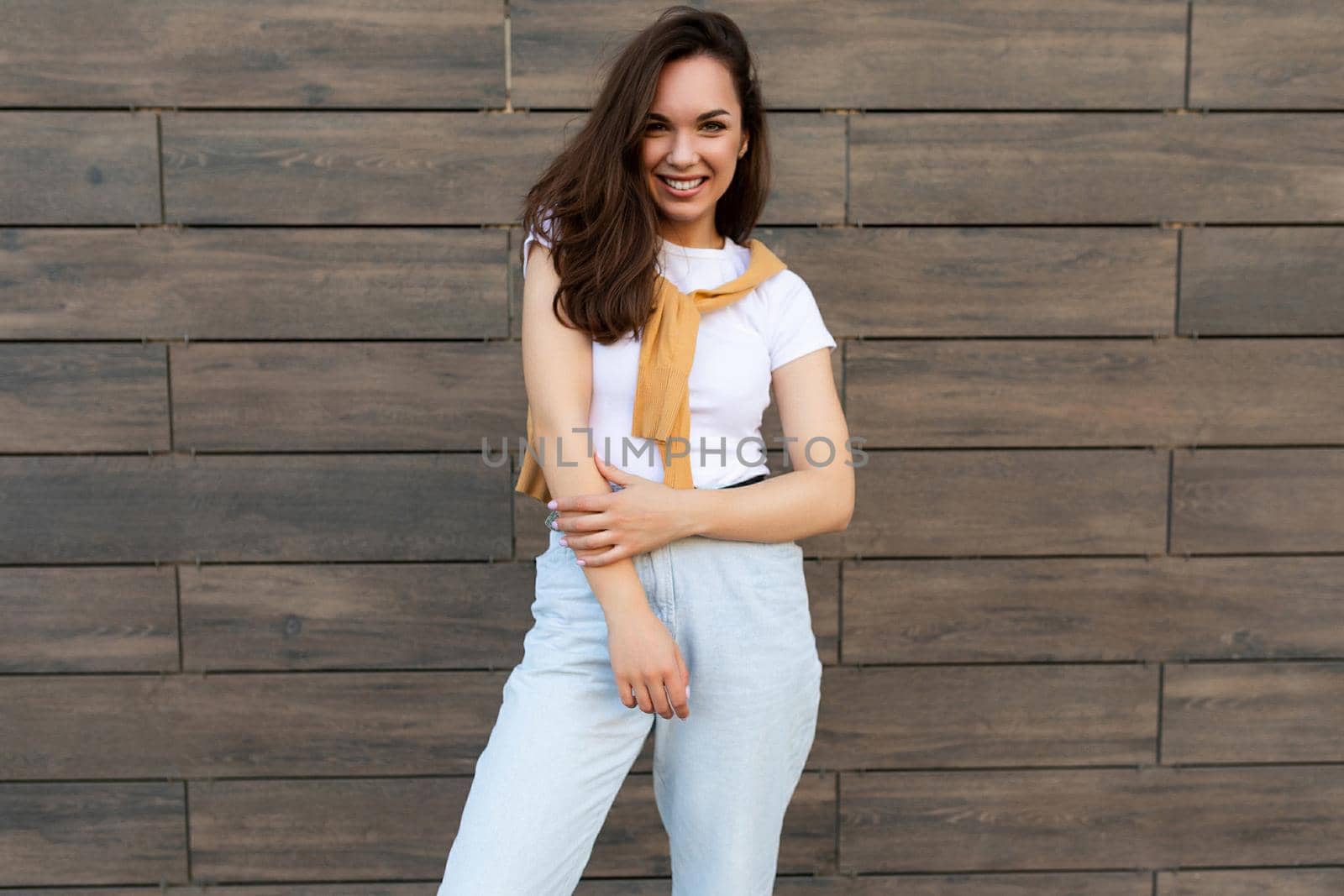 Photo of pretty attractive beautiful smiling happy positive charming young brunette young woman wearing stylish clothes: white t-shirt, yellow sweater and light blue jeans in the street near brown wall.