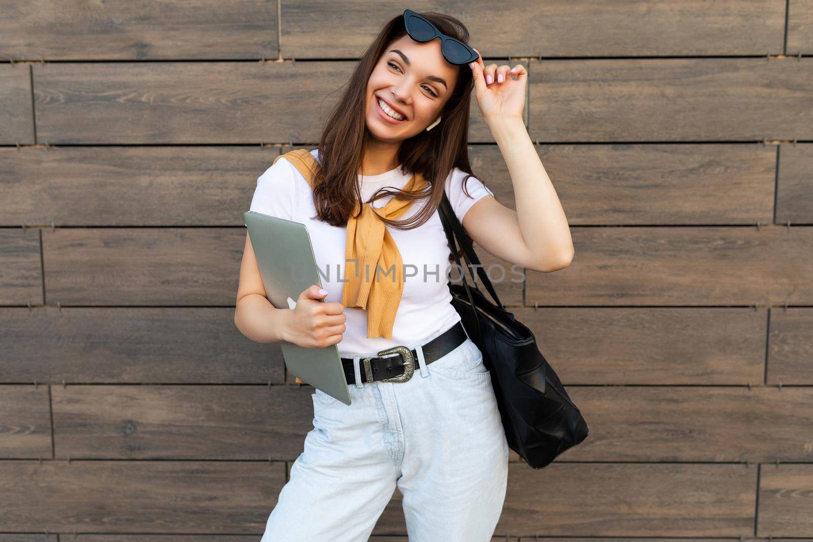 Photo of fascinating beautiful smiling happy positive charming young brunette girl wearing stylish clothes holding computer laptop and black sunglasses in white t-shirt and light blue jeans in the street near brown wall.