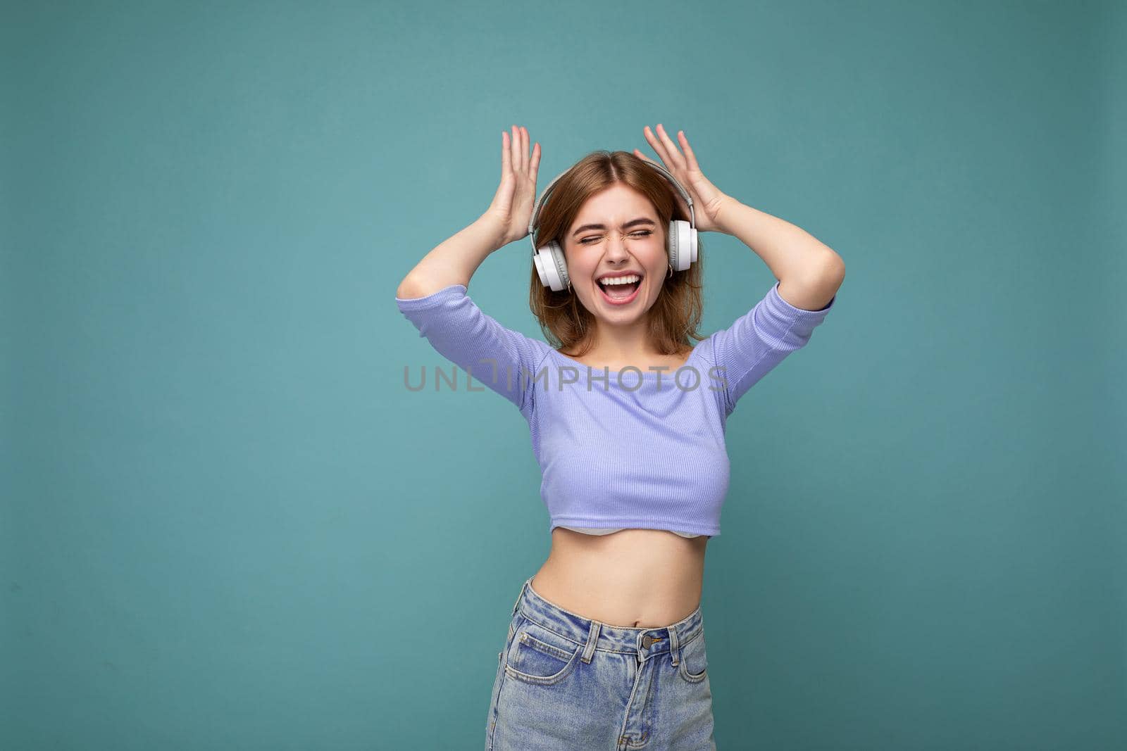 Charming happy smiling young blonde woman wearing blue crop top isolated on blue background with copy space for text wearing white wireless bluetooth headphones listening to good music dancing and having fun by TRMK