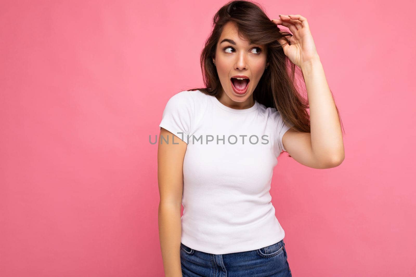 Photo portrait of young beautiful positive surprised amazed, shocked brunette woman in white t-shirt with mockup. Female person posing isolated near pink wall with empty space for text in studio.