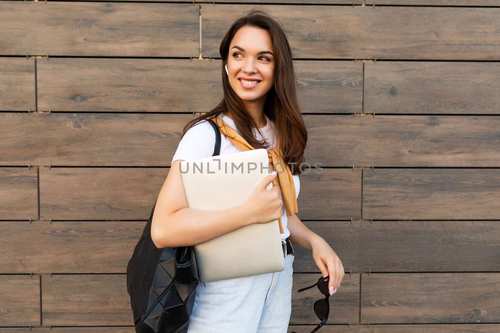 Photo of pretty attractive beautiful smiling happy positive charming young brunette young woman holding gray computer laptop wearing stylish clothes in white t-shirt and light blue jeans in the street near brown wall by TRMK