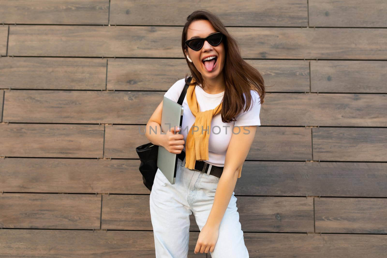 Photo of fascinating beautiful smiling charming young brunette girl wearing stylish clothes holding computer laptop and black sunglasses in white t-shirt and light blue jeans in the street near brown wall.