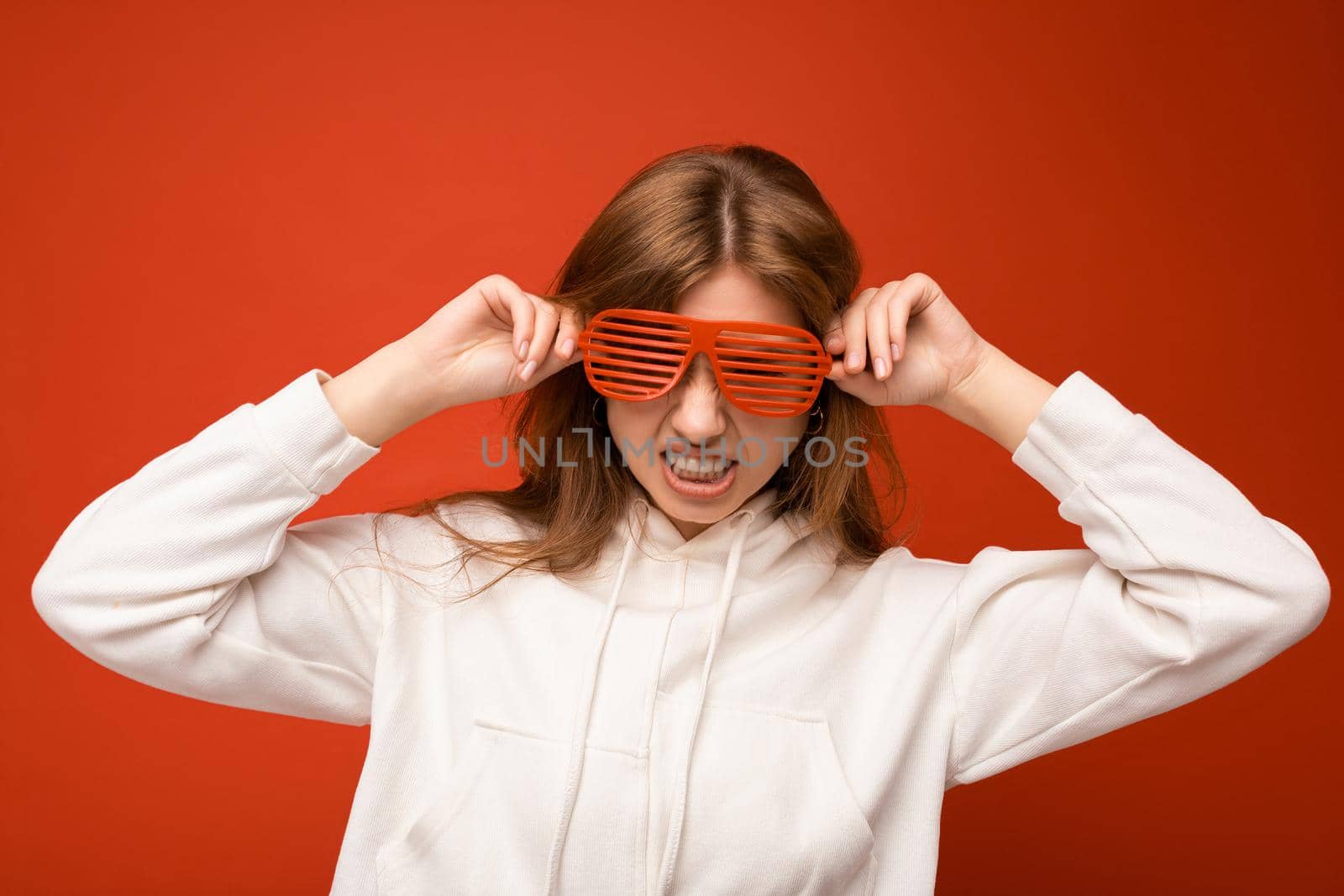 Photo shot of attractive positive happy young blonde woman isolated over colourful background with copy space for text wearing casual white hoodie and stylish glasses looking at camera and having fun.