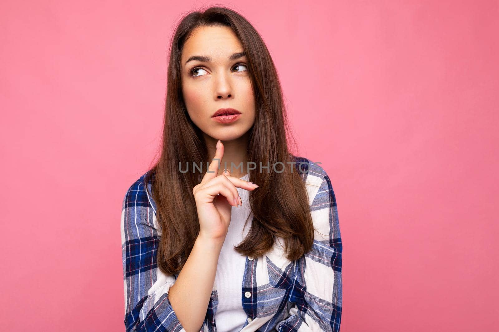 Photo portrait of young beautiful thinking and dreaming brunette woman in trendy blue and white shirt. Female person posing isolated near pink wall with empty space in studio.