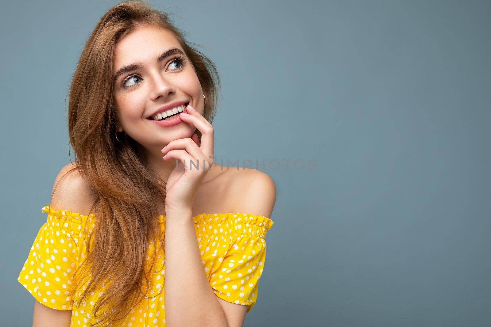 Close-up photo of young cute delightful beautiful attractive smiling positive happy dark blonde woman with sincere emotions isolated on blue background wall with copy space for text wearing stylish yellow dress. Positive concept.