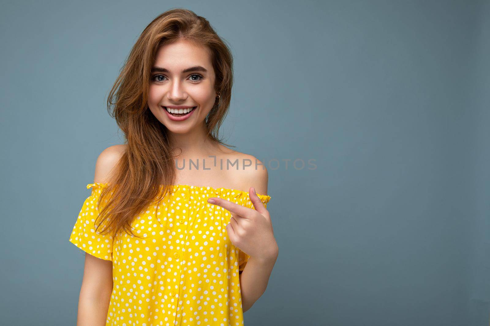 Photo of young cute delightful beautiful attractive smiling positive happy dark blonde woman with sincere emotions isolated on blue background wall with copy space for text wearing stylish yellow dress. Positive concept by TRMK