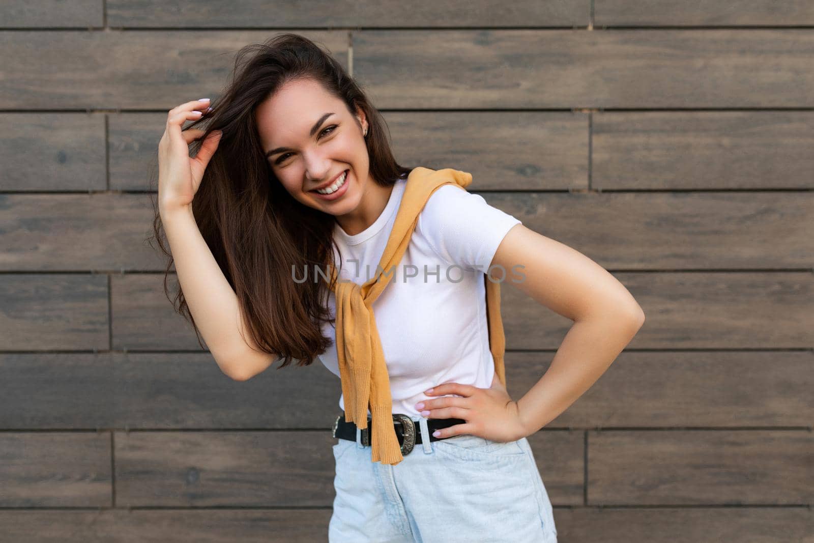 Photo of pretty attractive beautiful smiling happy positive charming young brunette young woman wearing stylish clothes: white t-shirt and light blue jeans in the street near brown wall.