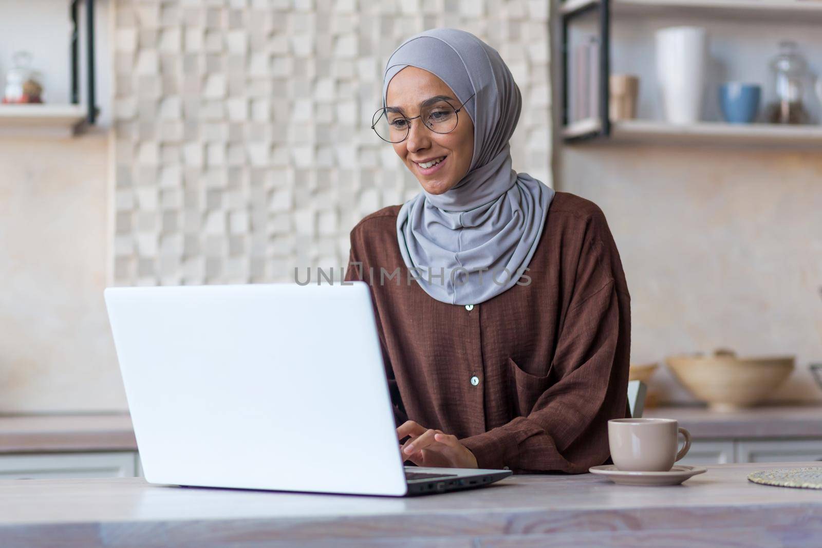 Young beautiful muslim woman in hijab and glasses studying remotely using laptop student woman in kitchen smiling and happy by voronaman