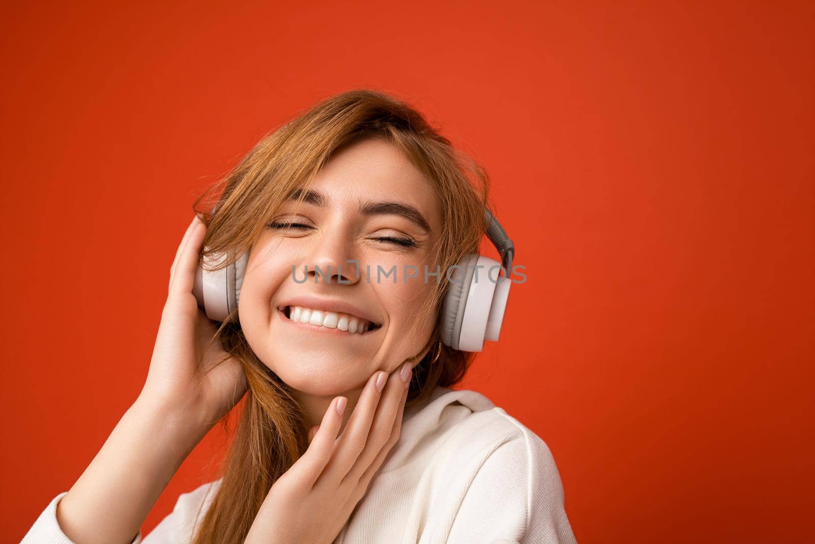 Close-up photo of beautiful happy smiling young blonde woman wearing white hoodie isolated over colourful background wall with copy space for text wearing white wireless bluetooth earphones listening to cool music and having fun.
